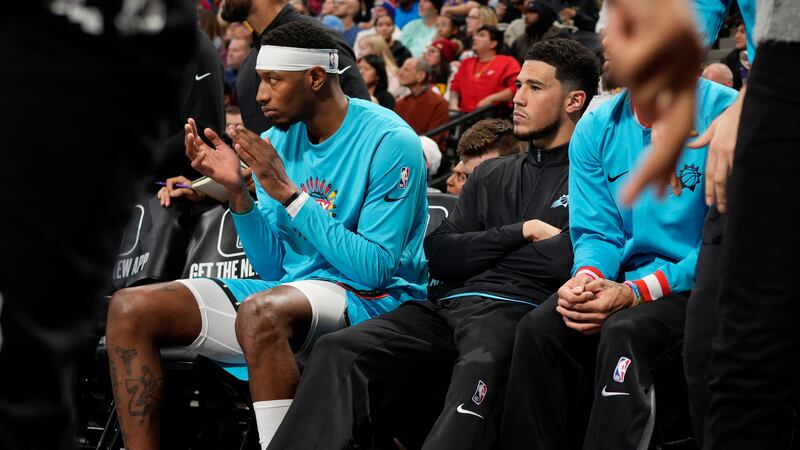 Phoenix Suns guard Devin Booker, wearing black, sits on the bench next to forward Torrey Craig...